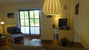Een gezellige woonkamer met een eettafel, rieten stoelen, een bank, een televisie en een groot raam met jaloezieën.