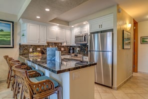 A kitchen with white cabinets, a stainless steel refrigerator, a microwave, a coffee maker, and a granite countertop. There are wooden chairs with blue cushions and a framed landscape painting on the wall.