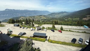 Un estacionamiento con varios autos, un edificio de techo inclinado y un paisaje montañoso al fondo.