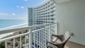 Un balcon agrémenté d’un fauteuil en osier, d’où l’on peut admirer une plage et un immeuble de grande hauteur doté de nombreux balcons.