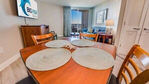 A dining area with a wooden table, four chairs, and two woven placemats. A television screen with 'BIG FISH Rentals' logo is mounted on the wall. - Coastal condo with balcony & beach access (Myrtle Beach)