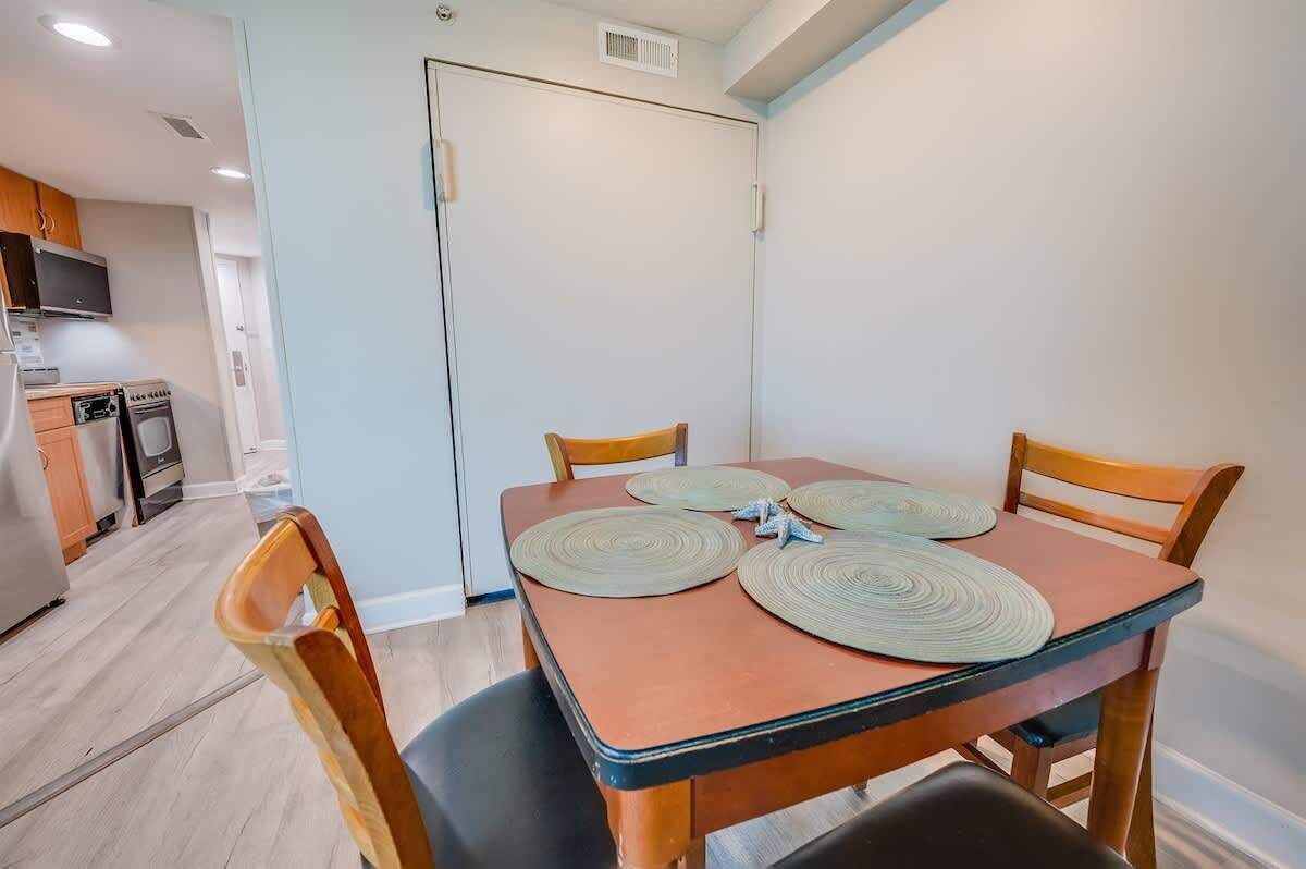 A dining area with a table set for four, surrounded by wooden chairs. A kitchen with appliances and cabinets is visible in the background.