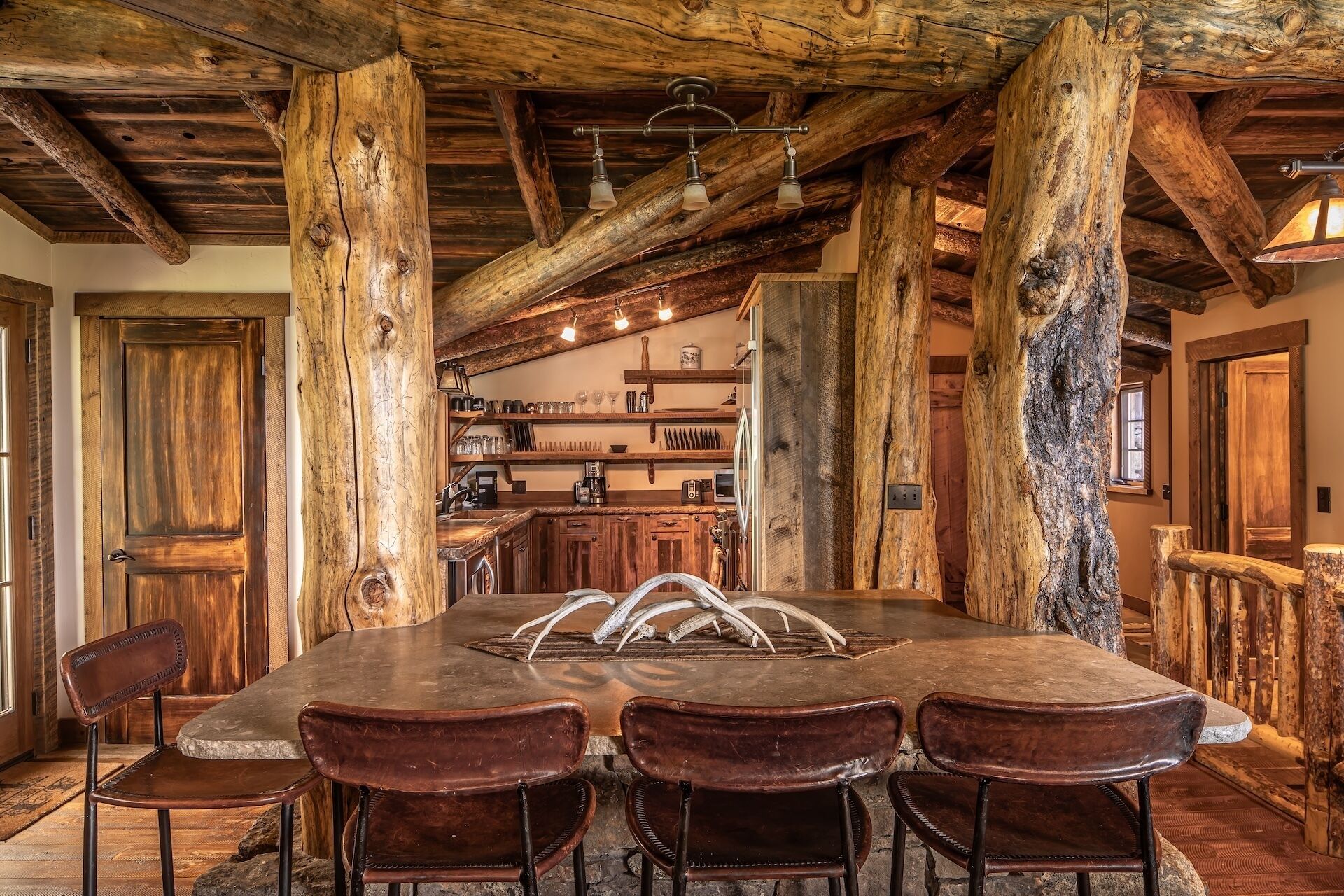 Un intérieur rustique avec une grande table en pierre et des chaises en cuir, des poutres en bois et un coin cuisine doté d’étagères.