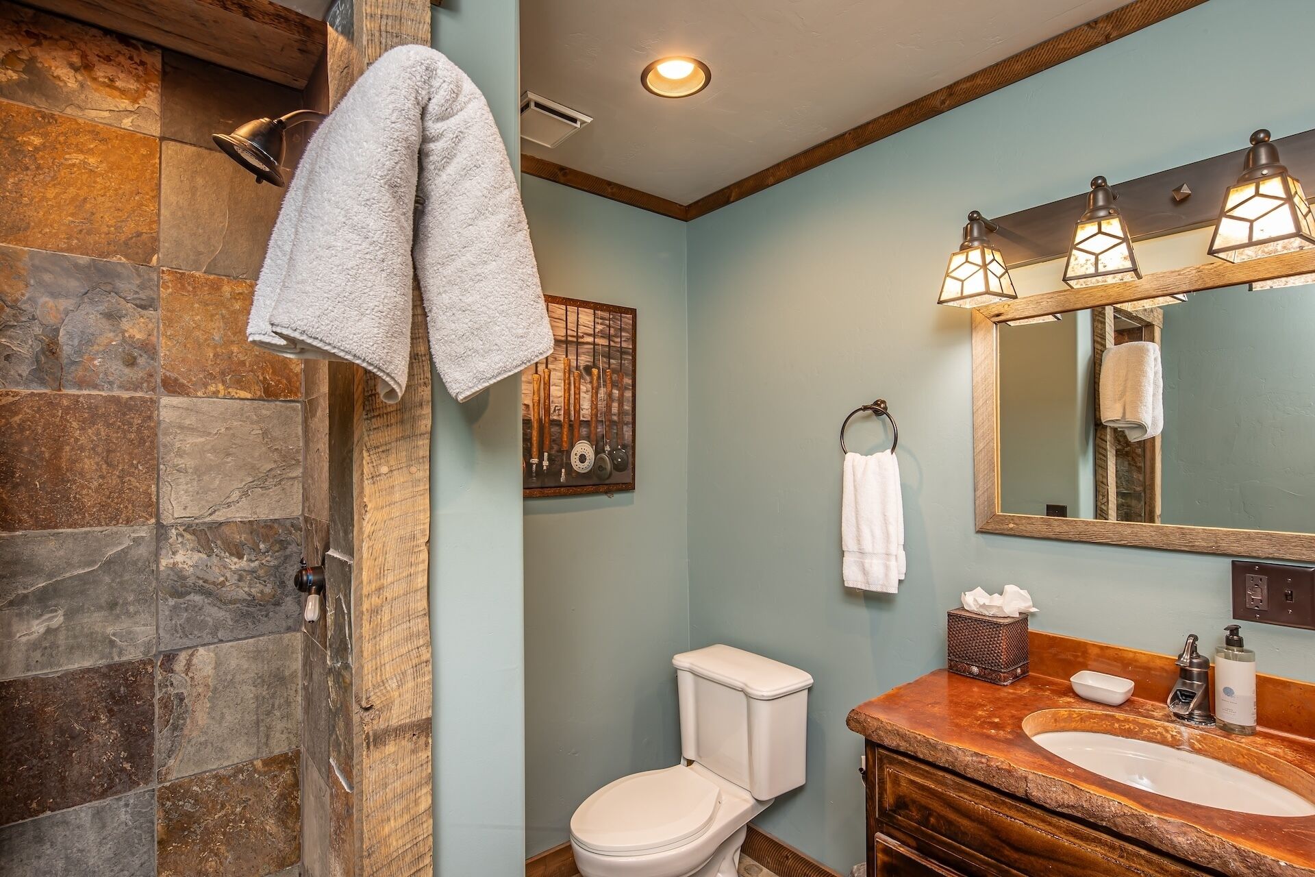 A bathroom with a stone shower enclosure, a toilet, a sink with a wooden countertop, and a mirror with three light fixtures.