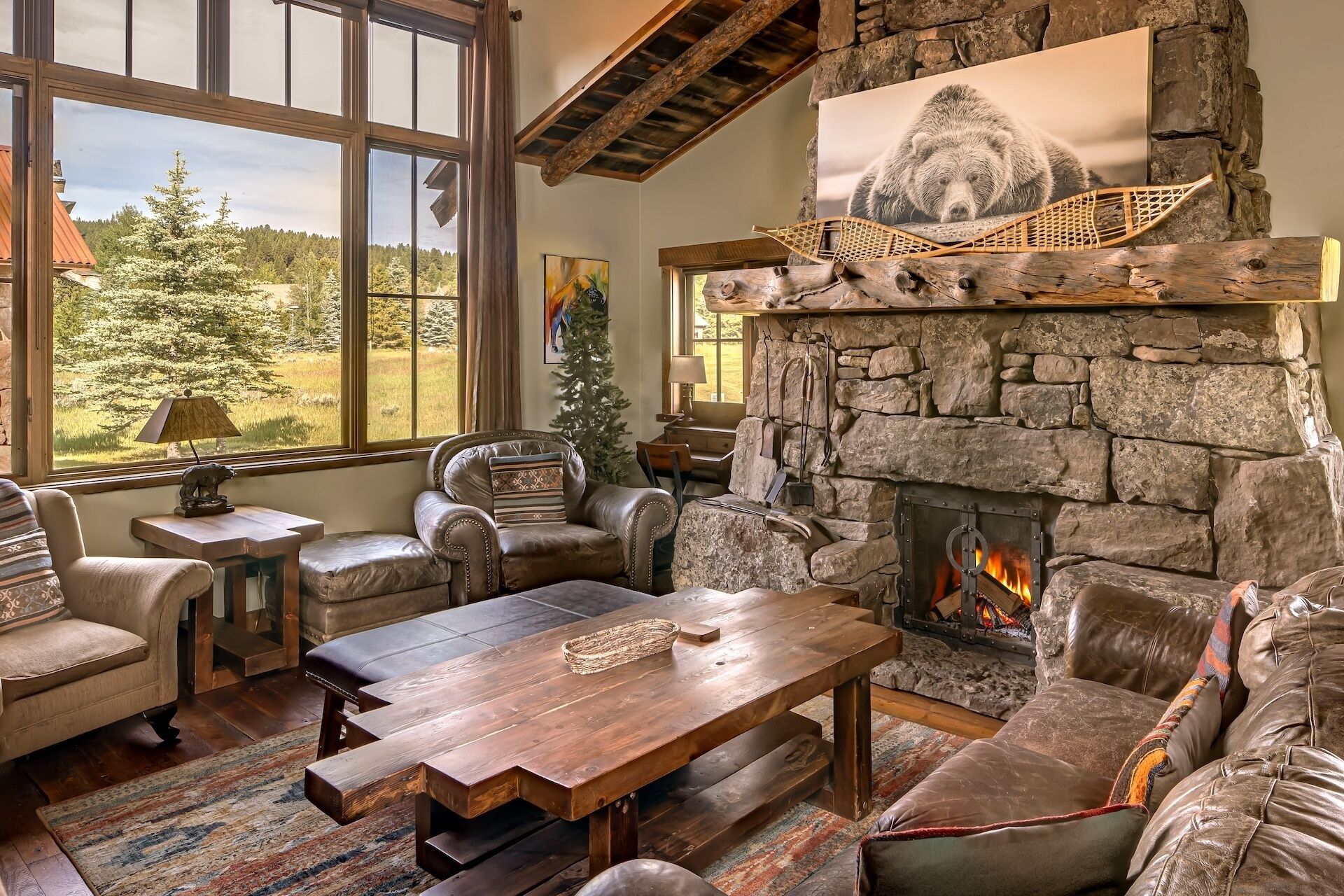 A cozy living room with a stone fireplace, leather furniture, and a view of a forest through large windows.