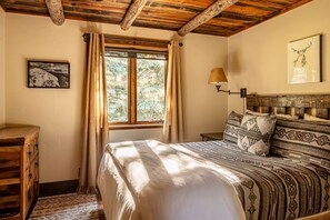 A room with a wooden ceiling, a bed with patterned bedding, a nightstand, a lamp, a window with curtains, and a framed picture on the wall.