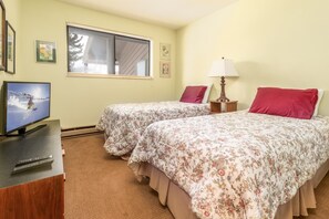 A hotel room with two beds, a TV, a lamp, and a window with a view of trees.