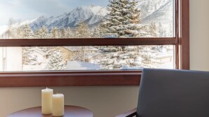 Een kamer met uitzicht op besneeuwde bergen door een raam. Er staat een klein, rond tafeltje met twee kaarsen en een enkele stoel.