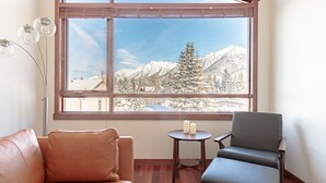 Een kamer met een groot raam dat uitzicht biedt op een besneeuwde berg, een leren bank, een donkere fauteuil en een kleine ronde tafel met kaarsen.