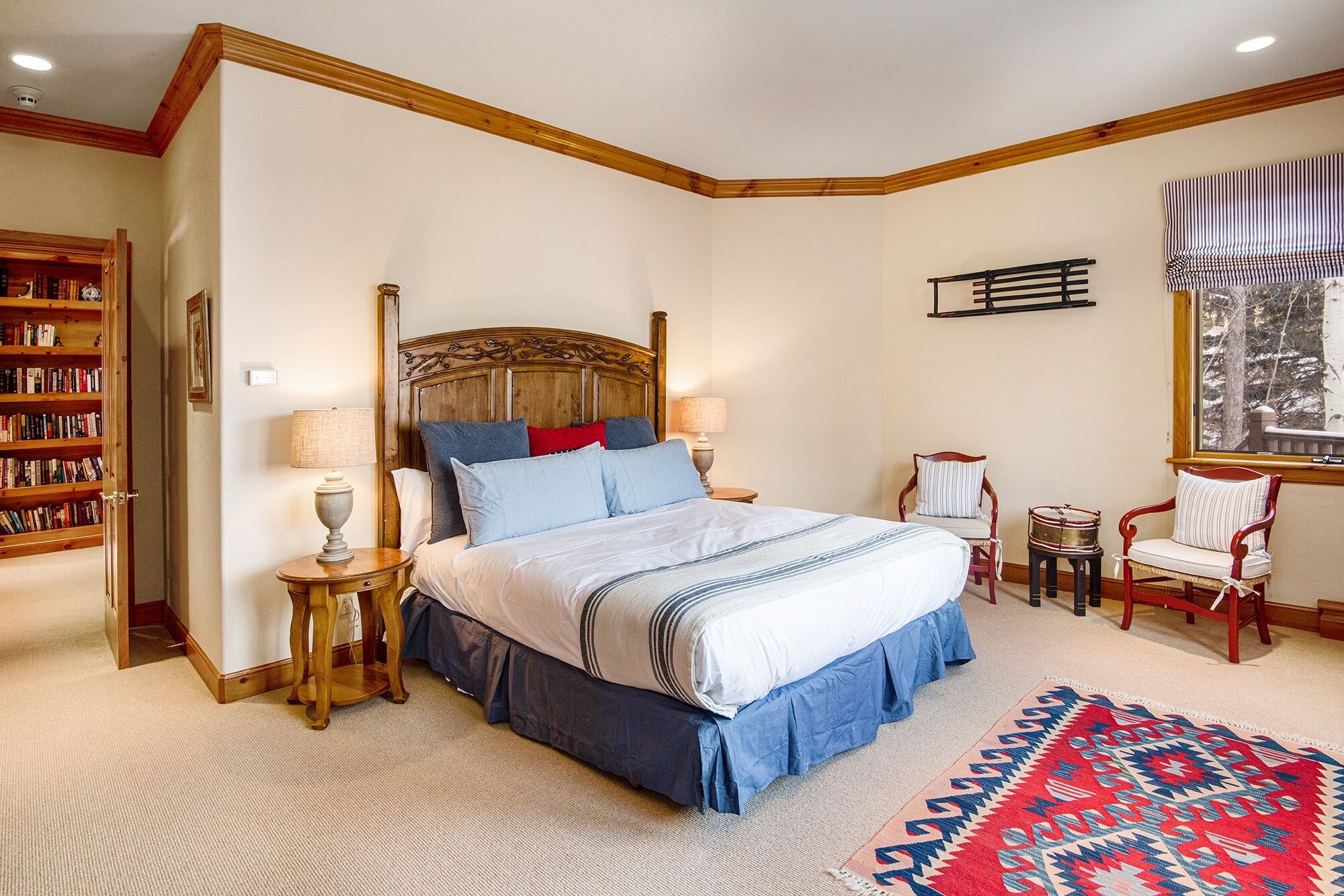 Quarto com uma cama grande, cabeceira de madeira, estante, pequena mesa com abajur, cadeira e janela com vista para árvores.