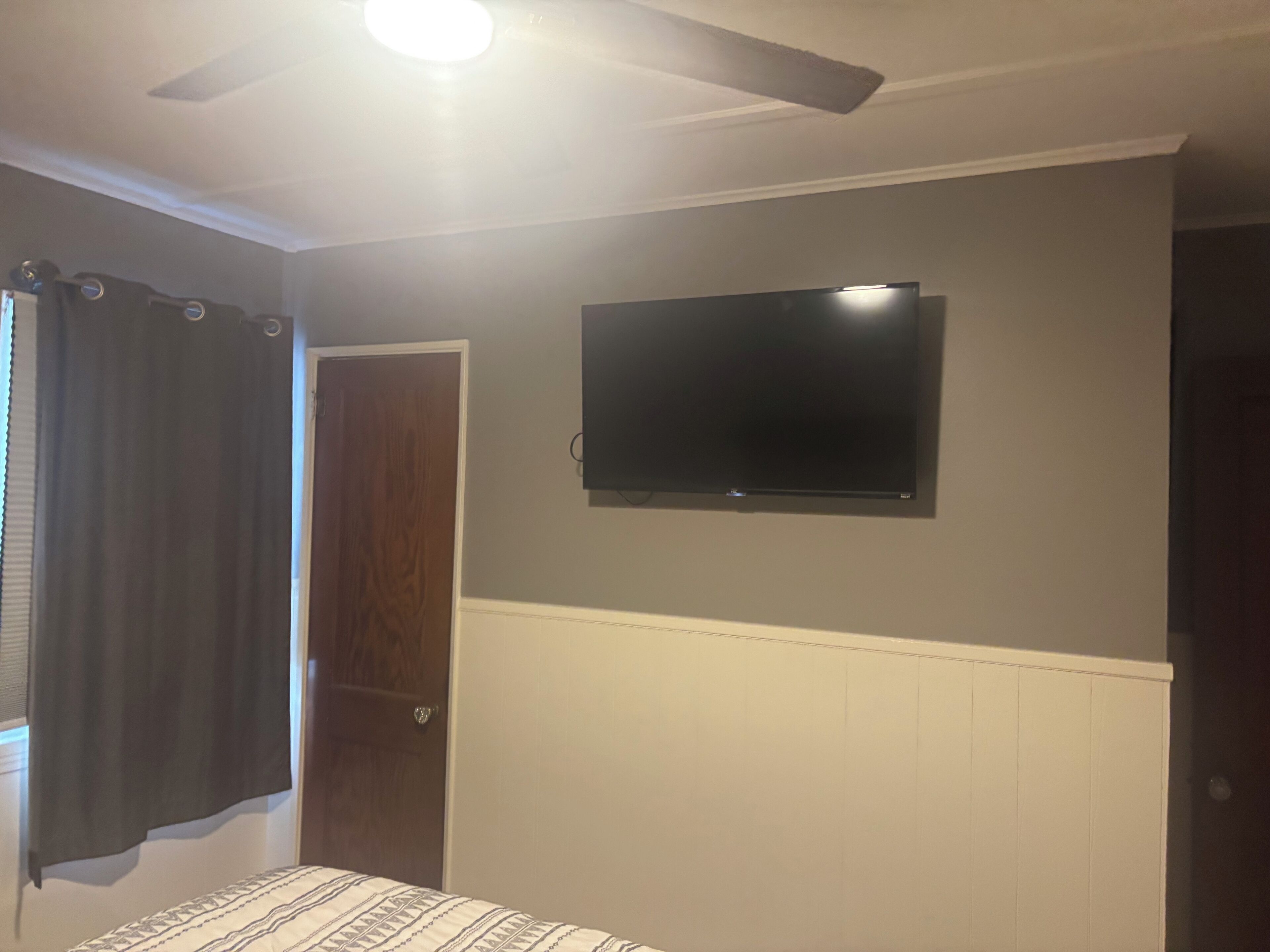A bedroom with a bed, a ceiling fan, a TV mounted on the wall, and a door.