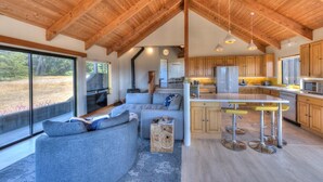 Une cuisine moderne avec des meubles en bois et un îlot central, un coin salon confortable avec un canapé et des coussins, et de grandes fenêtres offrant une vue sur l’extérieur.