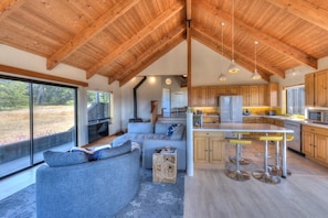 Una cocina moderna con armarios de madera y una isla central, un rincón de estar acogedor con un sofá y cojines, y ventanas amplias que ofrecen vistas al exterior.
