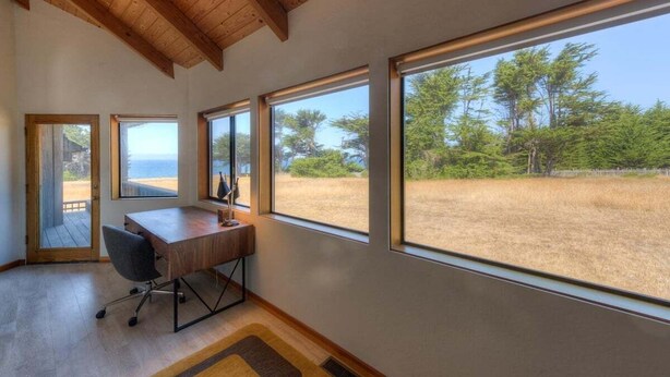 Une pièce avec un bureau en bois, une chaise et de grandes fenêtres donnant sur une plage et des arbres.