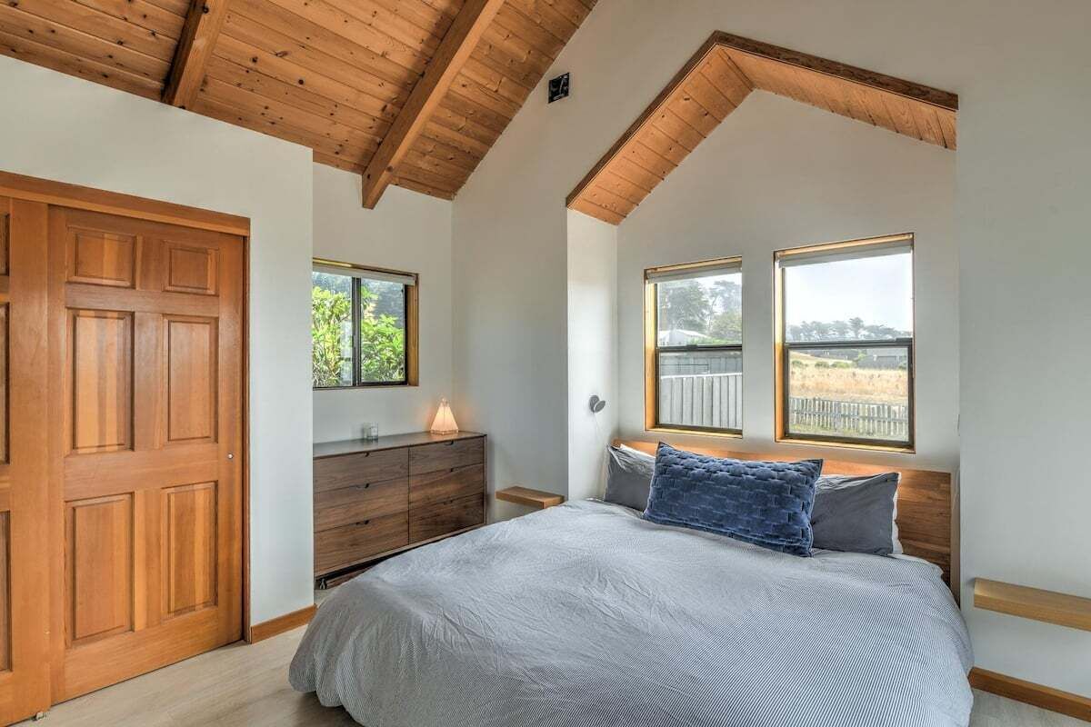 A bedroom with a wooden ceiling, a bed with grey bedding, a wooden dresser, and two windows with views.