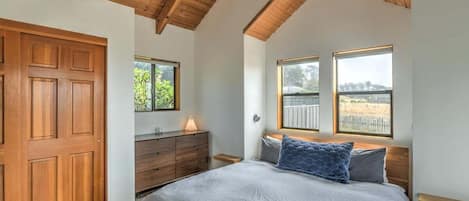 A bedroom with a wooden ceiling, a bed with grey bedding, a wooden dresser, and two windows with views.