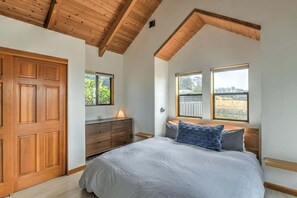 Quarto com teto de madeira, cama com roupa de cama cinza, cômoda de madeira e duas janelas com vista.