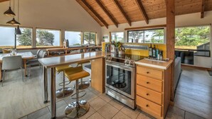 Une cuisine moderne dotée d’un îlot central, de poutres en bois, de grandes fenêtres et d’un coin repas avec des chaises.
