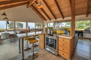 Una cocina moderna con una isla central, vigas de madera, ventanas amplias y un comedor con sillas.