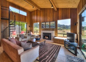 A spacious living room with a fireplace, comfortable seating, a television, and large windows offering a view of the outdoors.