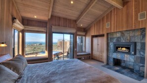 A bedroom with a large bed, a fireplace, and a view of the outdoors. - Berk | Ocean Views, Fireside Nights & Hot Tub (Sea Ranch)