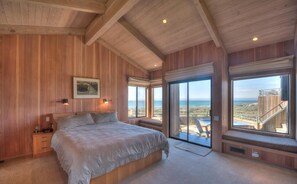 A bedroom with a large bed, wooden walls, a view of the beach, and a balcony with a railing. - Berk | Ocean Views, Fireside Nights & Hot Tub (Sea Ranch)