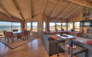A spacious living room with a large sofa, chairs, and a coffee table. There is a television and a fireplace. The room has large windows offering a view of the ocean. - Berk | Ocean Views, Fireside Nights & Hot Tub (Sea Ranch)