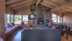 A cozy living room with a stone fireplace, a large gray sofa, and a television. - Berk | Ocean Views, Fireside Nights & Hot Tub (Sea Ranch)
