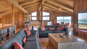 A spacious living area with a high ceiling, wooden beams, a large sofa set, a coffee table, and a bookshelf. - Berk | Ocean Views, Fireside Nights & Hot Tub (Sea Ranch)