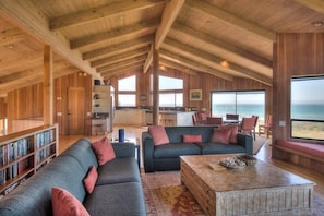 A spacious living area with a high ceiling, wooden beams, a large sofa set, a coffee table, and a bookshelf. - Berk | Ocean Views, Fireside Nights & Hot Tub (Sea Ranch)