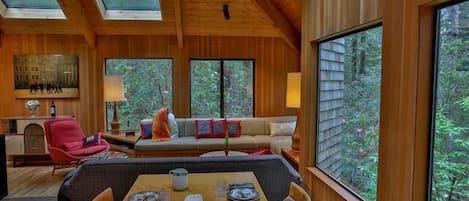 A wooden interior with a dining table set for four, a sofa, and a large window overlooking a forest.