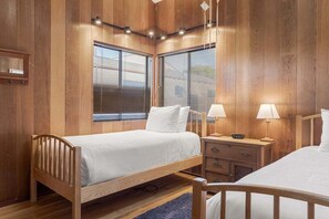 Um quarto com painéis de madeira, duas camas, uma mesa de cabeceira com luminárias e uma janela com persianas.