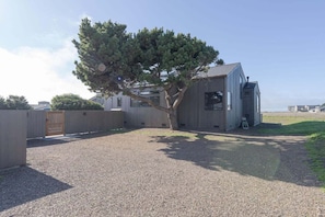 A modern building with a gravel driveway, a wooden fence, and a tree. - Gray Whale | Hot Tub Nights & Meadow Views (The Sea Ranch)