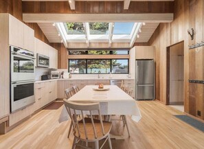 A modern kitchen with a dining area, wooden cabinetry, and a skylight. - Gray Whale | Hot Tub Nights & Meadow Views (The Sea Ranch)
