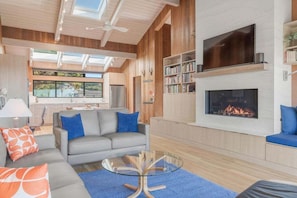 A modern living room with a fireplace, a sofa, a coffee table, and a bookshelf. - Gray Whale | Hot Tub Nights & Meadow Views (The Sea Ranch)