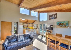 Ein geräumiges Wohnzimmer mit einer Ledersofa, einem Holzessttisch und Stühlen, eine Küche im Hintergrund und ein großes Fenster, das natürliches Licht hereinlässt.