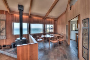 A wooden interior with a large window overlooking the sea, a fireplace, and a dining area.