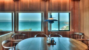 Un coin repas avec une table et des chaises en bois, de grandes fenêtres donnant sur l’océan et un luminaire suspendu.