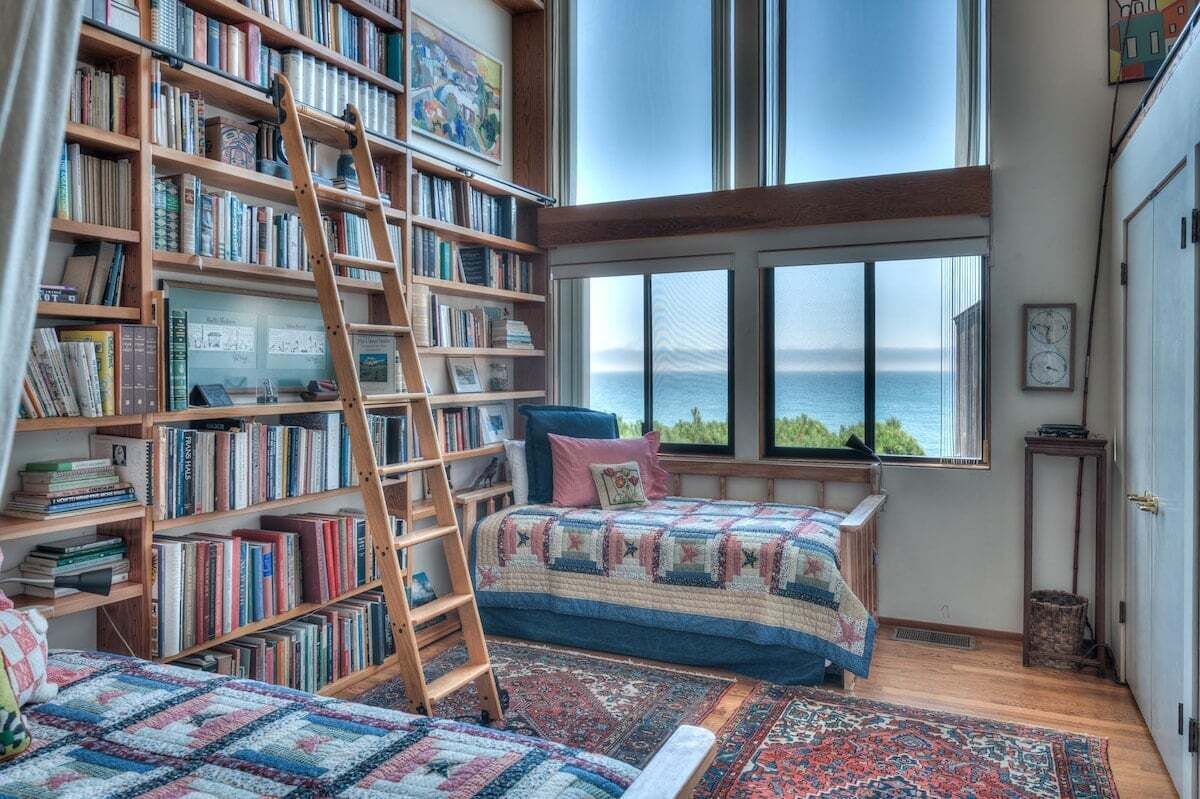 A room with a large bookshelf, a ladder, a bed with a patterned quilt, and a view of the sea through a large window.