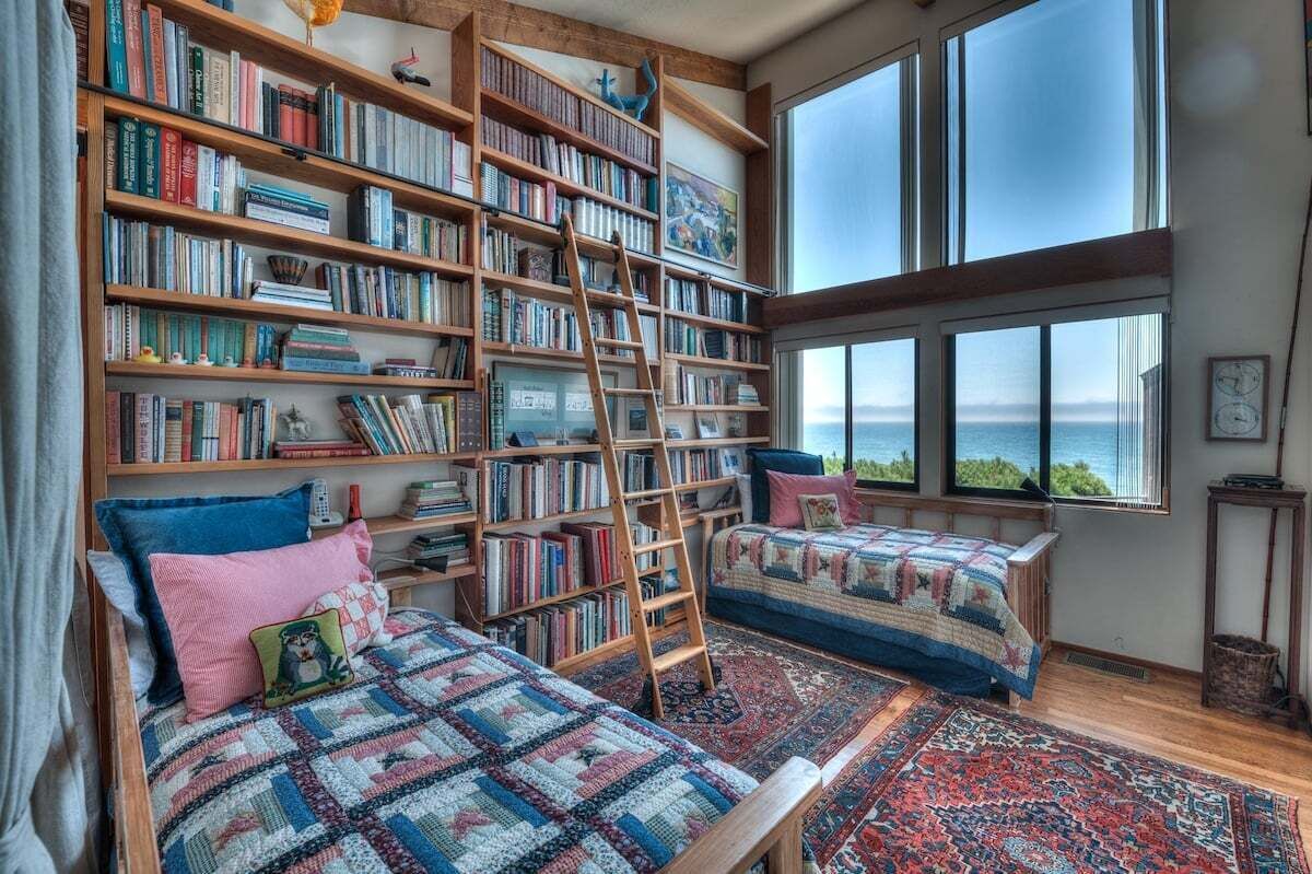 Une chambre avec une grande bibliothèque, une échelle, un lit recouvert d’une couvre-lit à motifs et une vue sur la mer par de grandes fenêtres.