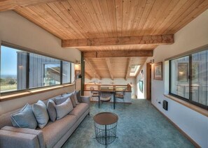 A spacious room with a wooden ceiling, a sofa with cushions, a round table, and large windows offering a view. - Sandpiper Nest | Coastal Escape Near Walk On Beach (The Sea Ranch)
