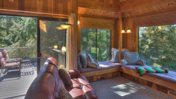 A wooden cabin interior with a large window, a cushioned bench, and a seating area.