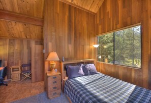 A wooden cabin bedroom with a bed, bedside lamps, a desk, and a window with a view of trees.