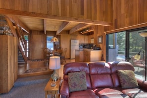 A wooden interior with a leather sofa, dining area, kitchen, and large windows.