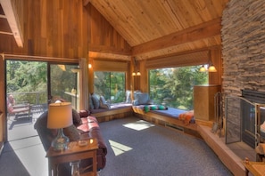 A cozy cabin interior with a stone wall, wooden ceiling, and large windows offering a view of the forest.