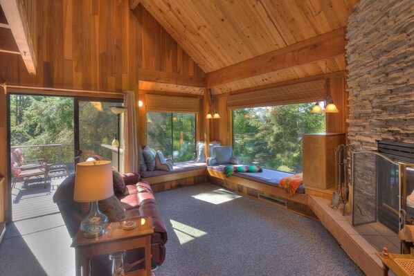 A cozy cabin interior with a stone wall, wooden ceiling, and large windows offering a view of the forest.