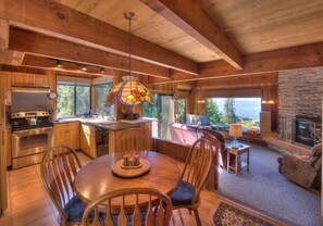 A spacious kitchen with wooden cabinets, a dining area with a round table and chairs, and a stone fireplace. - Second Wind | Coastal Bliss & Hot Tub (The Sea Ranch)