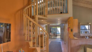 A spacious living area with a wooden spiral staircase, a television, and a kitchen area with a wooden counter. - Silver Mist | Pool with Stargazing Decks (The Sea Ranch)