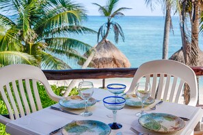 Tavolo apparecchiato per due con piatti, bicchieri e posate, con vista su una spiaggia, palme e l'oceano.