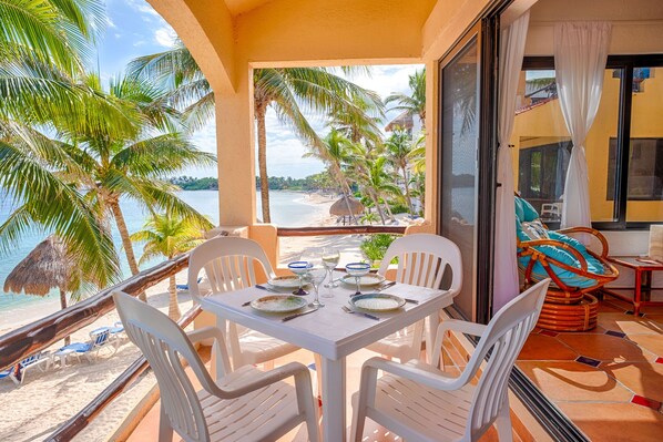 Un balcone con un tavolo apparecchiato per due, con vista su una spiaggia, palme e una capanna con tetto di paglia.